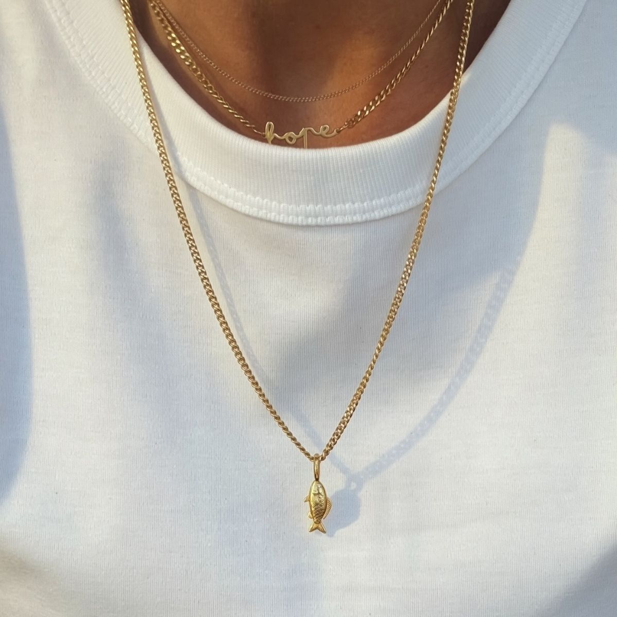 Halsband med en guldfärgad fiskberlock hängande från en kedja. Berlocken har detaljer som visar fiskens form och textur. Ögonen har en vit cz-sten.